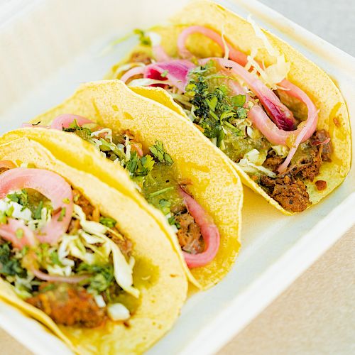 The image shows three tacos in a takeout container, filled with meat, onions, cilantro, and other toppings, on a beige background.