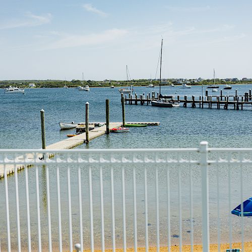 A serene waterfront scene with docks, sailboats, a white fence, and a blue umbrella on the beach under a clear sky, ending the sentence.