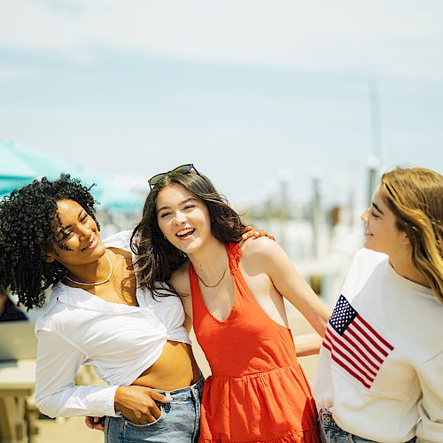 Three women are smiling and posing together outdoors near a waterfront, with tables and umbrellas in the background.