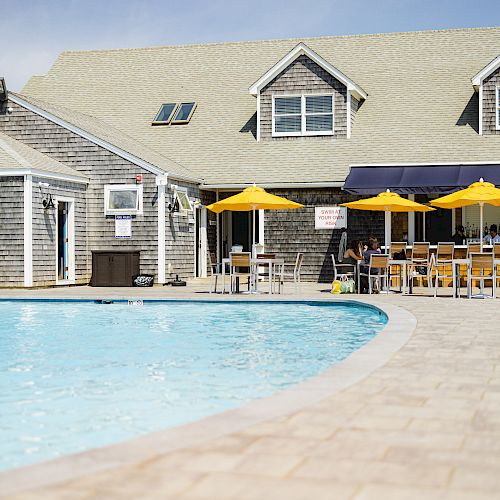 A swimming pool next to a building with outdoor seating and yellow umbrellas, with lounge chairs lined up along the poolside.