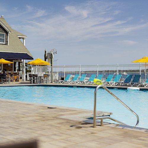 A swimming pool area with yellow umbrellas, lounge chairs, and a nearby building under a clear blue sky.