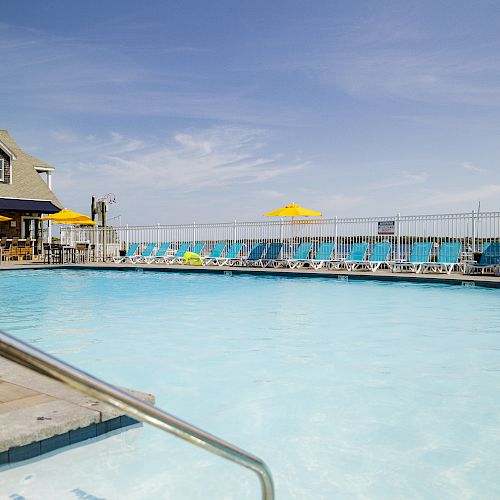 A swimming pool with a nearby building, lounge chairs, and umbrellas positioned around it under a clear sky.