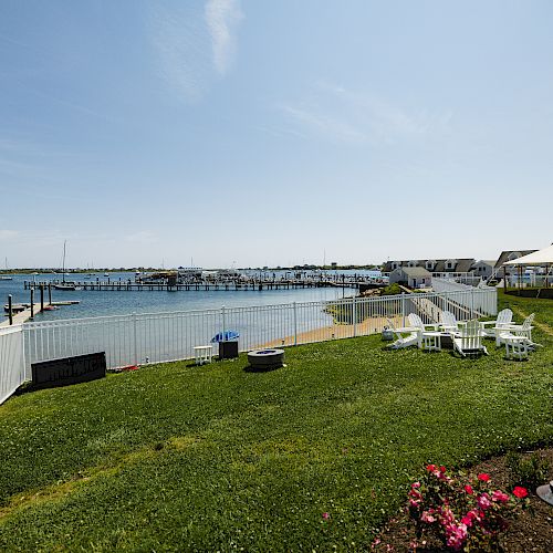 A scenic waterfront view with green grass, white fences, chairs, a tent, a dock, and calm water under a clear sky ending the sentence.