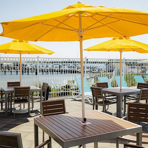 An outdoor seating area with wooden tables and chairs under yellow umbrellas, beside a pool with lounge chairs and a fence in the background.