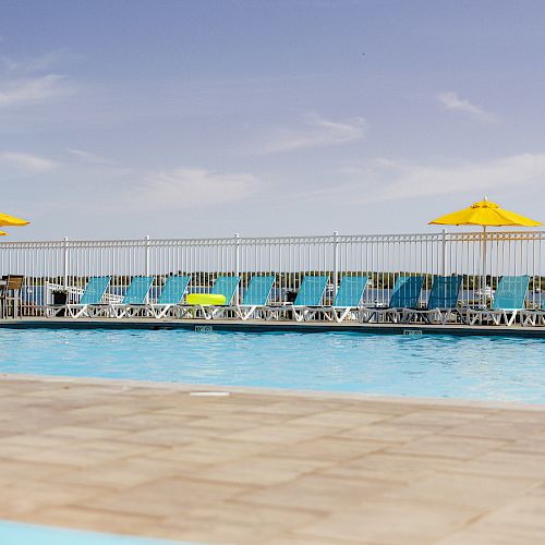 A swimming pool with lounge chairs and yellow umbrellas on a sunny day, surrounded by a metal fence with a clear blue sky in the background.