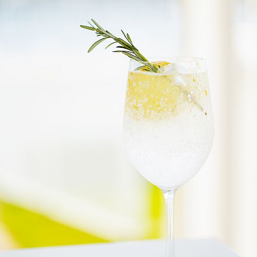 A refreshing beverage in a wine glass with ice, garnished with a sprig of rosemary and a lemon twist, placed on a smooth surface.