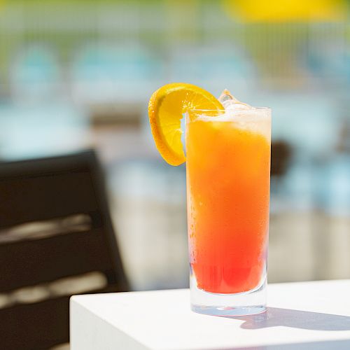 A tall, colorful cocktail garnished with an orange slice sits on a white table, with blurred out-of-focus chairs and pool area in the background.