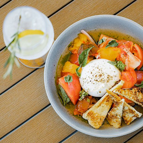 A bowl with tomatoes, basil, sliced bread, and a poached egg on a wooden table; a glass of water with lemon is beside it.