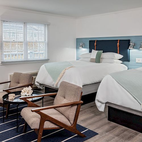 A modern hotel room with two beds, two chairs with a small table between them, and a floor lamp. The beds have white linens and light blue accents.