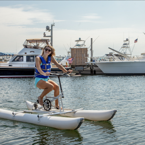 A person wearing a life jacket rides a water bike in a marina with several boats docked in the background.