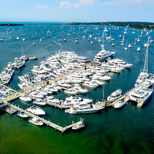 An aerial view of a marina filled with numerous yachts and boats docked at piers, with many more boats anchored in the surrounding waters.