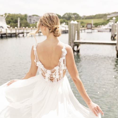 A woman in a white, backless dress is standing on a dock by the water, her hair styled in an updo, looking away from the camera.