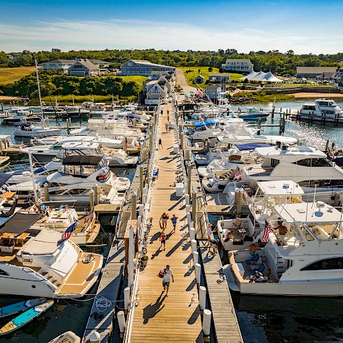 A dock filled with various boats and yachts, with people walking along the pier, under a clear blue sky, with buildings and greenery in the background.