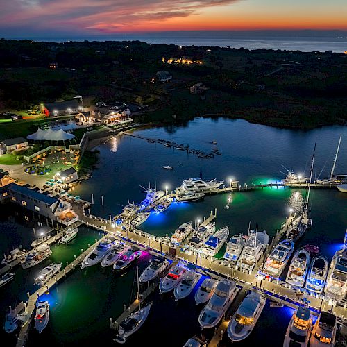 Aerial view of a marina at dusk with boats docked; lit with colorful lights, surrounded by water, buildings, and a scenic sunset.