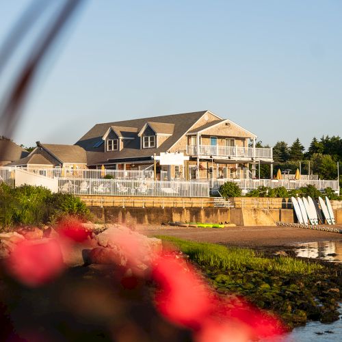 A waterfront property with a large house, paddleboards, and kayaks on the shore, with blurred flowers in the foreground.