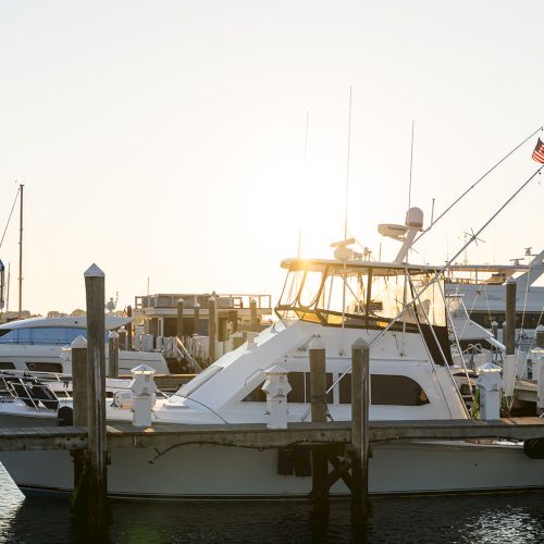 A white yacht is docked at a marina during sunset, with a building and more boats visible in the background.