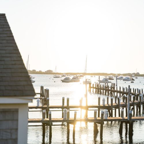 The image shows a serene waterfront scene at sunset with a dock and boats anchored in the water, and a building with a shingled roof in the foreground.