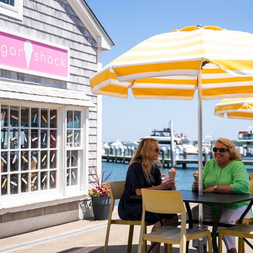 Two women sit at a table under yellow-striped umbrellas near a building with a sign reading 