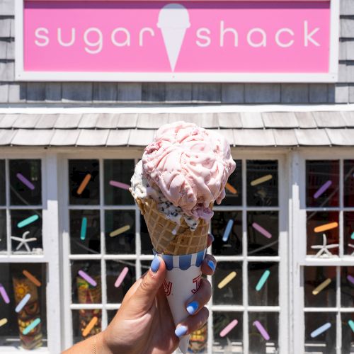 A hand holds a waffle cone with two scoops of ice cream in front of a shop with a pink sign that reads 