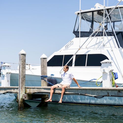 A person sitting on a dock near a boat, talking on the phone, with feet dangling over the water, under a clear blue sky.