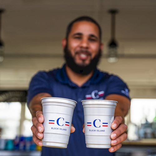 A person is holding two cups towards the camera, smiling, likely promoting a brand or business. The background appears to be a café or bar.