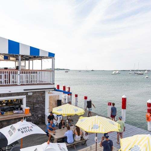 People sitting under umbrellas at a waterside restaurant overlooking a marina with boats in the distance, on a clear day.