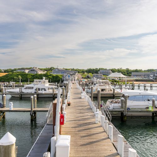 A marina with several boats docked along wooden piers extends into the water under a partly cloudy sky, with buildings and greenery in the background.