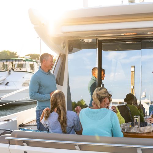 A group of people enjoying a meal on a yacht, docked at a marina on a sunny day, with boats and greenery visible in the background.