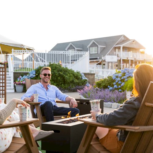 A group of people is relaxing on wooden chairs around a fire pit in an outdoor setting, with a house and tent in the background.