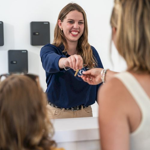 A smiling woman behind a counter hands keys to another woman with a child. It appears to be a reception desk or check-in counter.