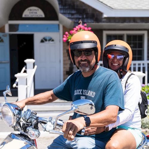 Two people are riding a scooter in front of a building, both wearing helmets and sunglasses, with flowers in the background and greenery around.