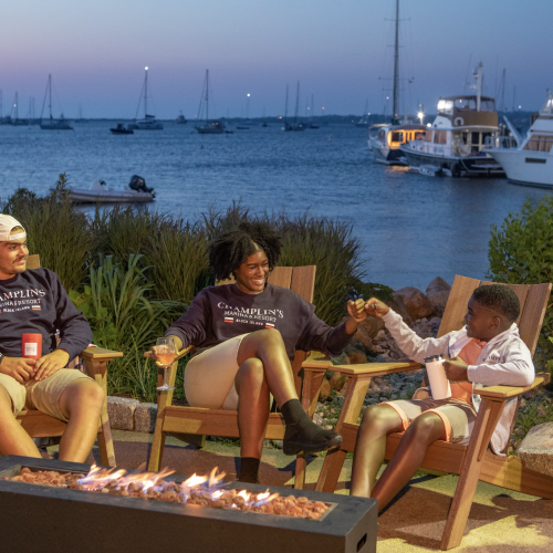 Three people are sitting around a fire pit near a waterfront, with boats docked in the background.