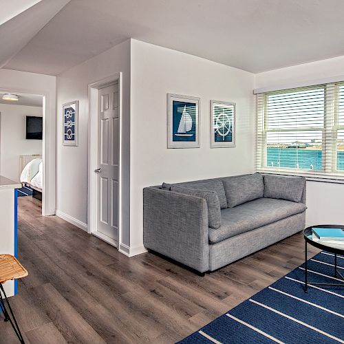 A modern, well-lit living room with a grey couch, white wall art, large window, kitchenette, bar stool, and a small round coffee table with a book.