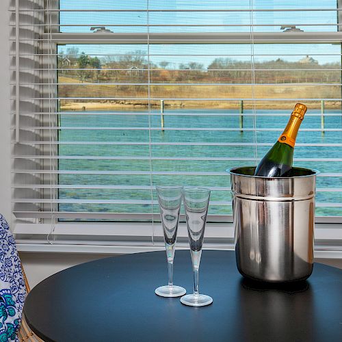 A table by a window with a bottle of champagne in an ice bucket, two flutes, a hat, and a piece of fabric. Water scenery is visible outside.