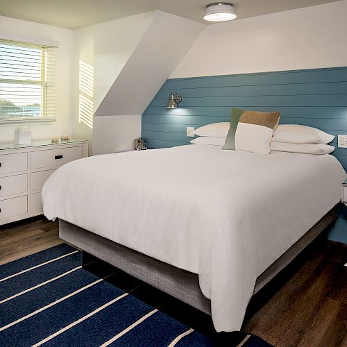 A cozy bedroom with a white bed, blue accent wall, striped rug, bedside tables, lamps, and a chest of drawers under a window with blinds.