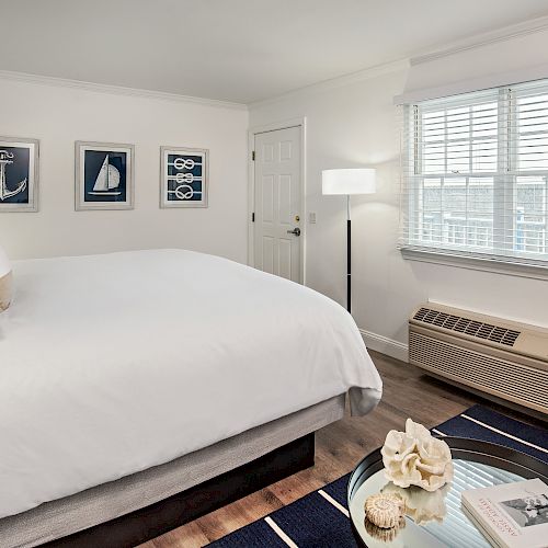 A cozy bedroom featuring a large bed, nautical-themed artwork, a window with blinds, a chair, a table with decor, and various light fixtures.