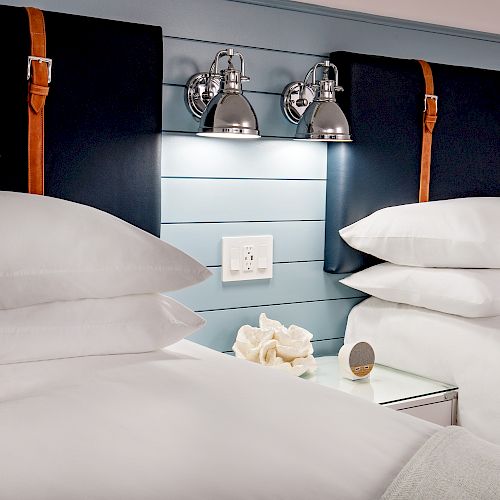 This image shows two neatly made beds with white linens, pillows, a small table in between, wall-mounted reading lights, and a seashell decoration.