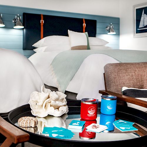 A cozy hotel room features two beds, nautical-themed decor, a small round table with snacks and games, and two armchairs with cushions.