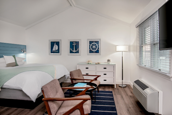 A cozy bedroom with a nautical theme, featuring a bed, chairs, dresser, wall art, floor lamp, striped rug, window, and an air conditioner.