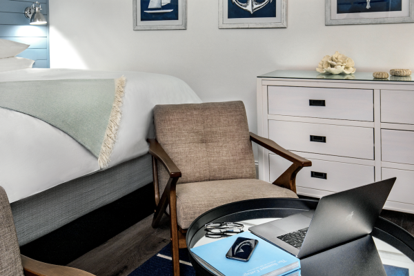 A cozy bedroom with a bed, a chair, a dresser, nautical-themed wall art, and a table holding a laptop, phone, glasses, and books.