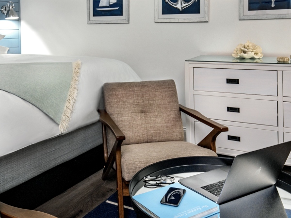 A cozy bedroom with a bed, a chair, a dresser, nautical-themed wall art, and a table holding a laptop, phone, glasses, and books.
