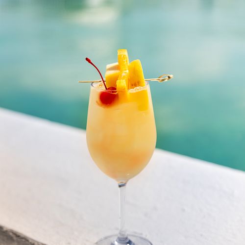 A tropical drink garnished with pineapple slices and a cherry is served in a wine glass by a poolside, against a blurred water background.