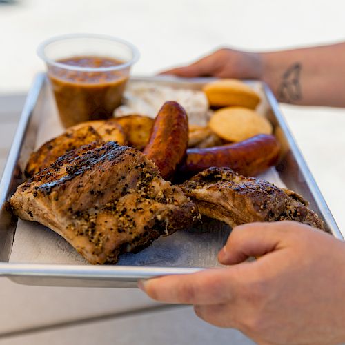 A person holding a tray of food with grilled meats, including ribs, sausage, a side of beans, bread, and an additional side cup.