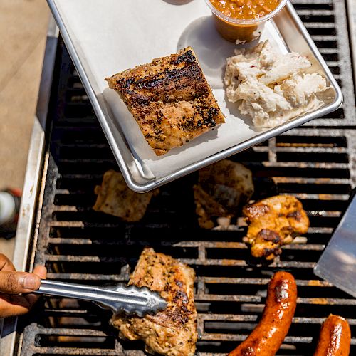 The image shows a barbecue with grilled sausages and chicken, alongside a tray containing cornbread, coleslaw, baked beans, and a piece of seasoned meat.