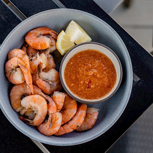 The image shows a bowl of cooked shrimp with a lemon wedge and a small bowl of cocktail sauce, placed on an outdoor table.