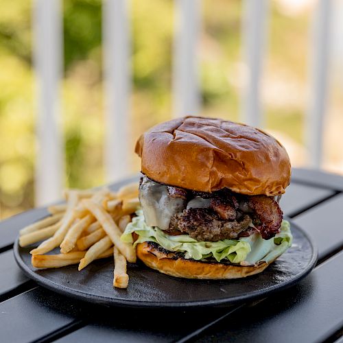 A delicious burger with lettuce and bacon on a plate accompanied by a serving of French fries, set on an outdoor table.