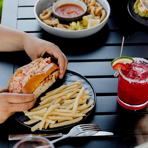 A person holding a sandwich next to a plate of fries and a red cocktail, with additional plates of food on a dark tabletop.