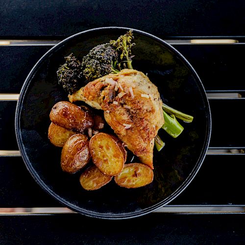 A black plate with roasted chicken, baby potatoes, and green vegetables is placed on a black surface with slats.