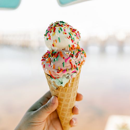 A hand holds an ice cream cone with two scoops topped with colorful sprinkles, against a blurred background of water and bridge structures.