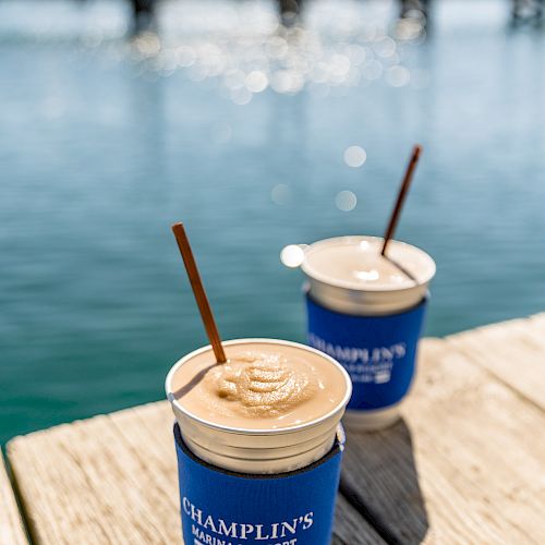 Two beverages in blue Champlin's Marina cups with straws are on a wooden surface by the water, with a dock and calm sea in the background.